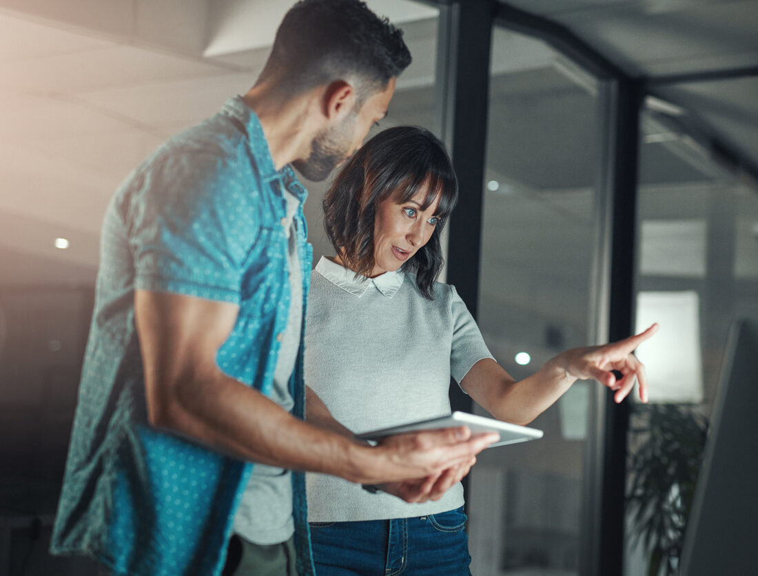 cropped shot of two businesspeople standing together in the office at night and having a discussion while using technology